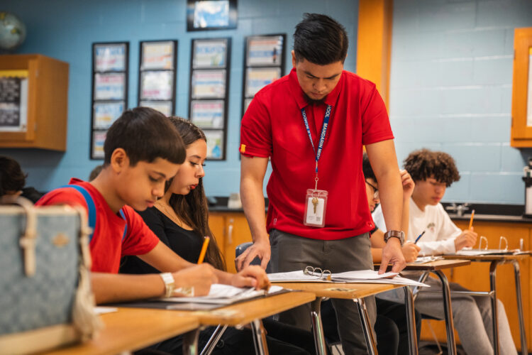 A middle school teacher checks the work of his students at their desk.